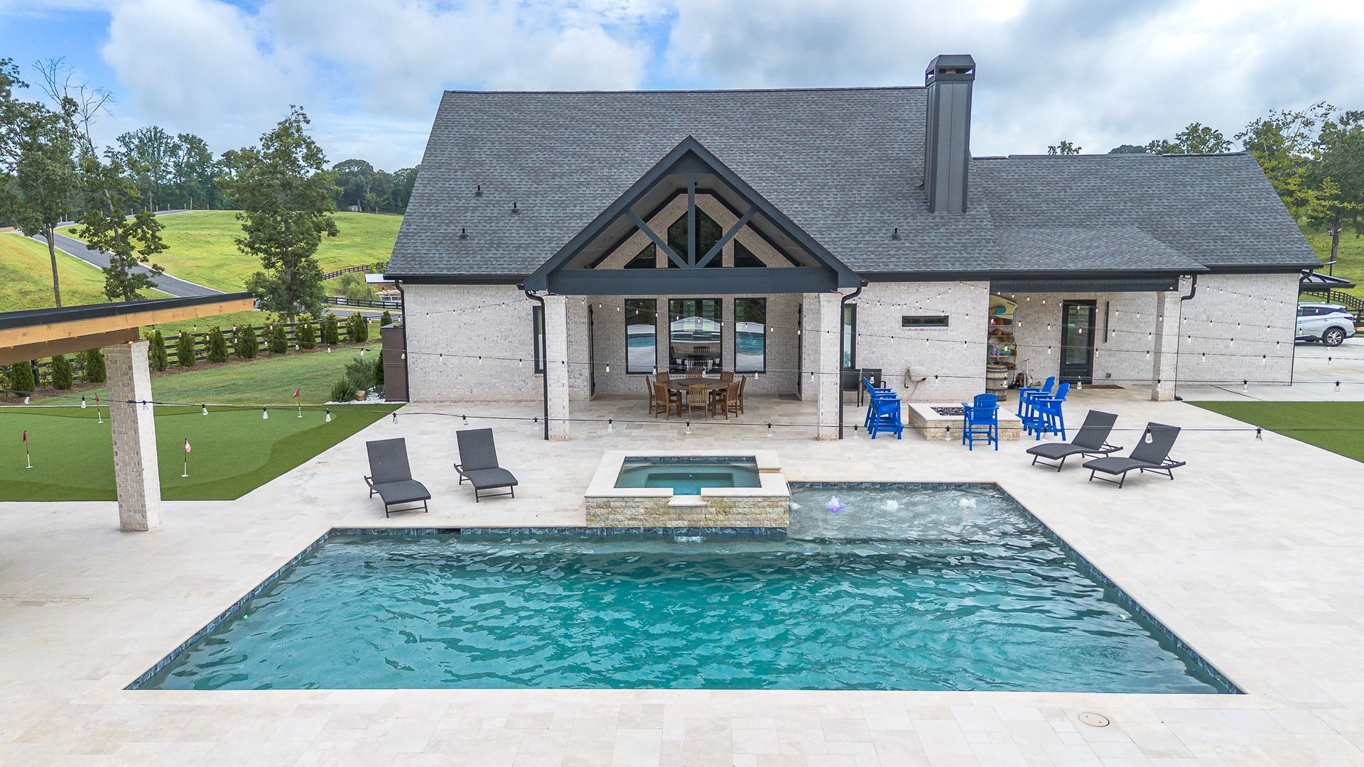 House with pool, spa, and outdoor seating. Grey roof, white brick, and lush green landscape.