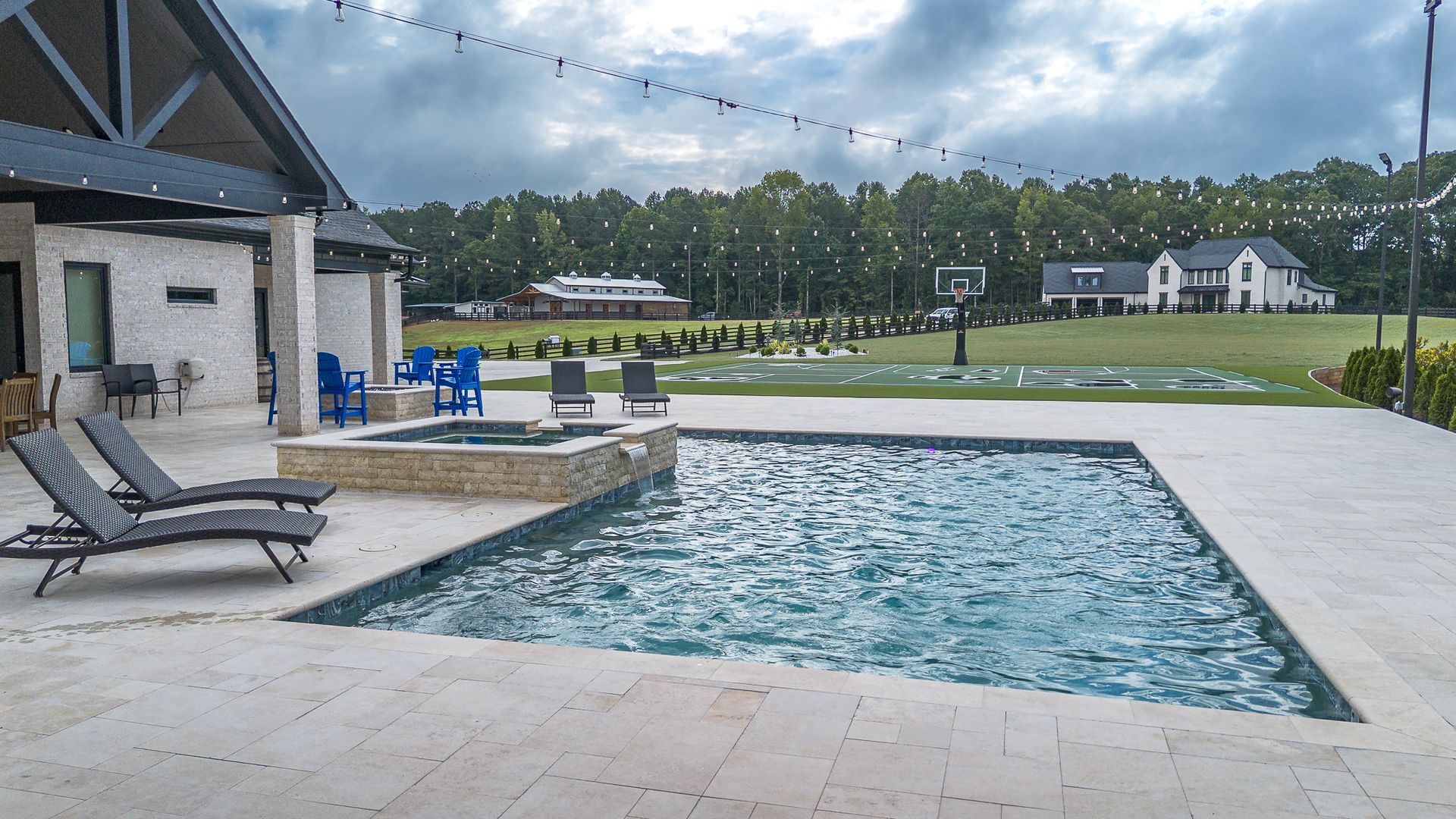 Outdoor pool and hot tub in a backyard setting with a modern home, trees, and cloudy sky.