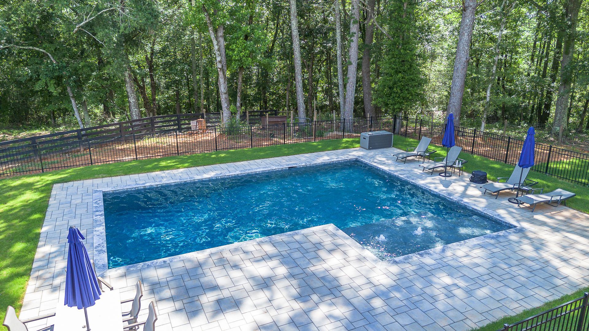 Pool with blue water, grey patio, lounge chairs, and blue umbrellas, surrounded by trees and a black fence.