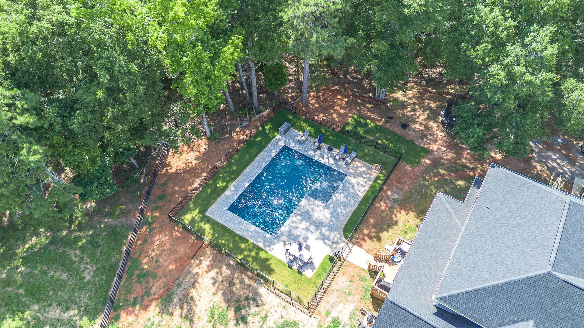 Aerial view of a blue rectangular pool surrounded by grass and trees, next to a house with a gray roof.