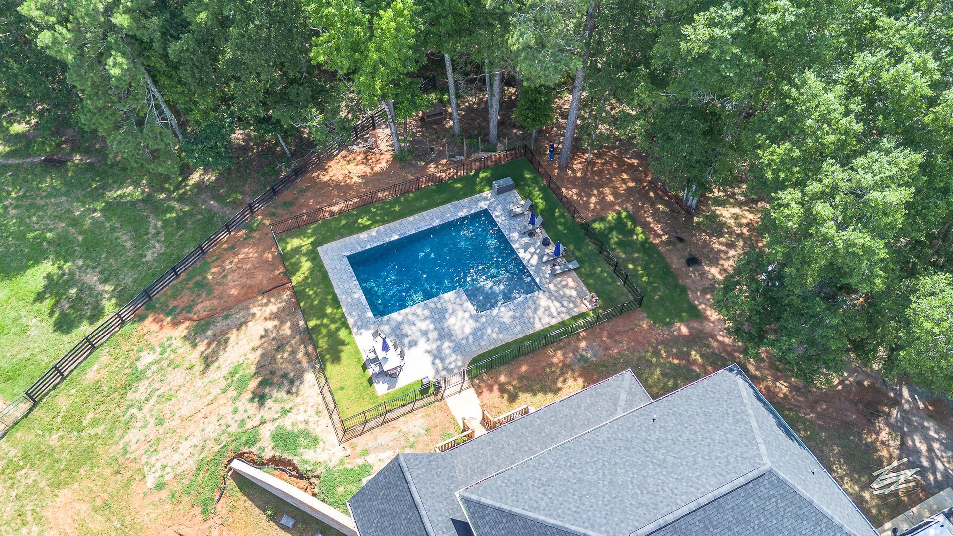 Aerial view of a rectangular pool with a gray patio, surrounded by a lawn, trees, and a house.