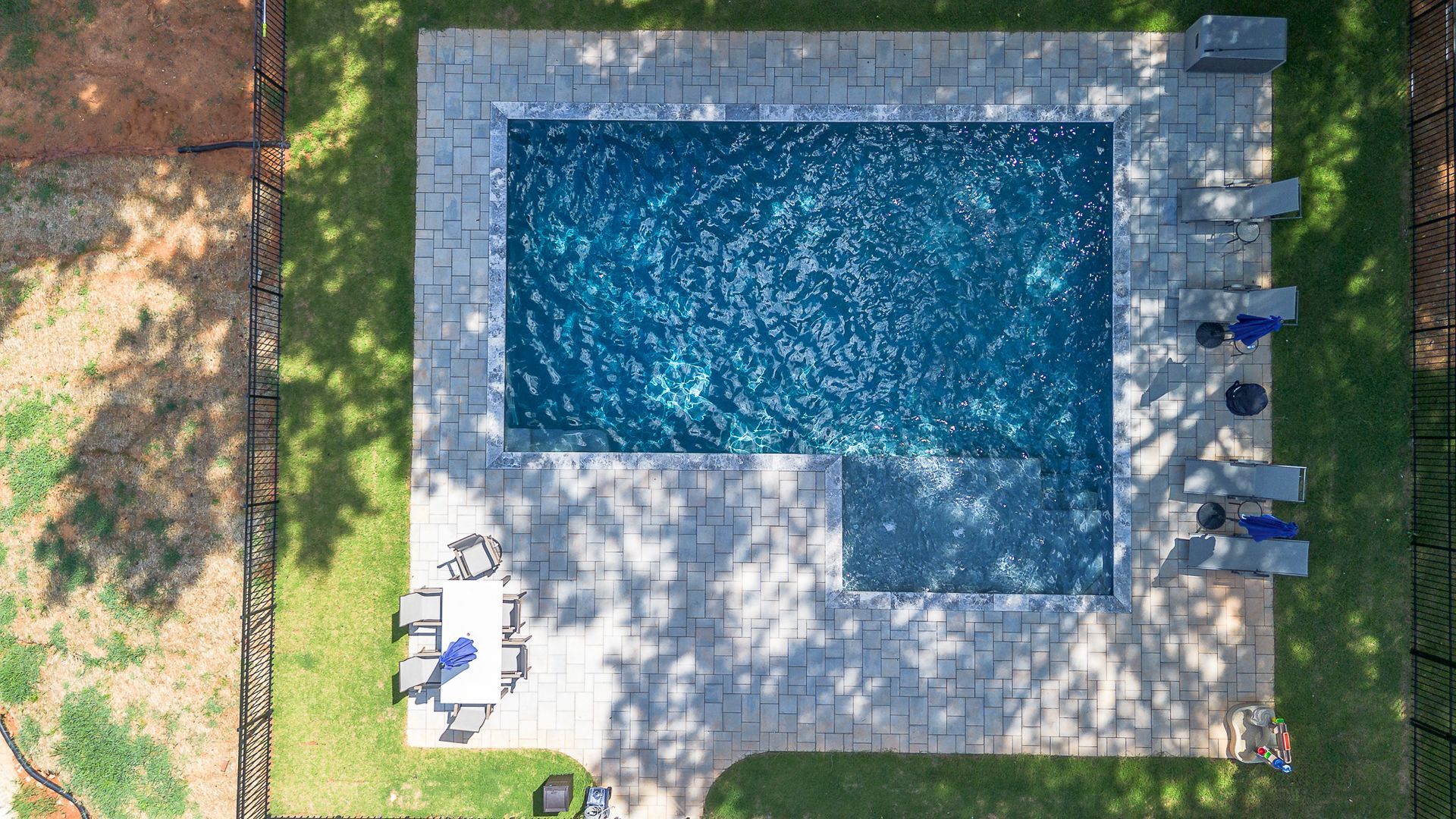 Overhead view of a rectangular swimming pool with blue water surrounded by a paved patio and green grass.