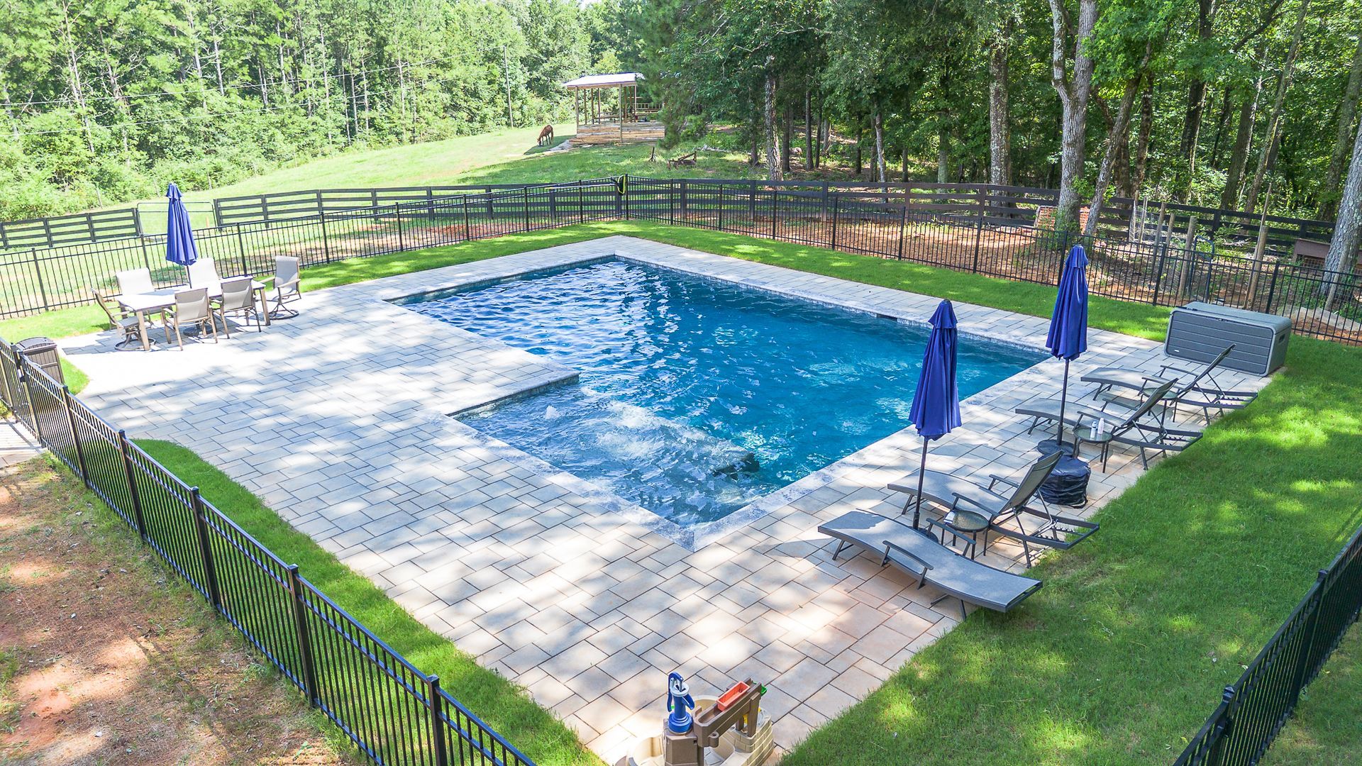 Swimming pool surrounded by a black fence, patio furniture, and a grassy yard with trees in the background.