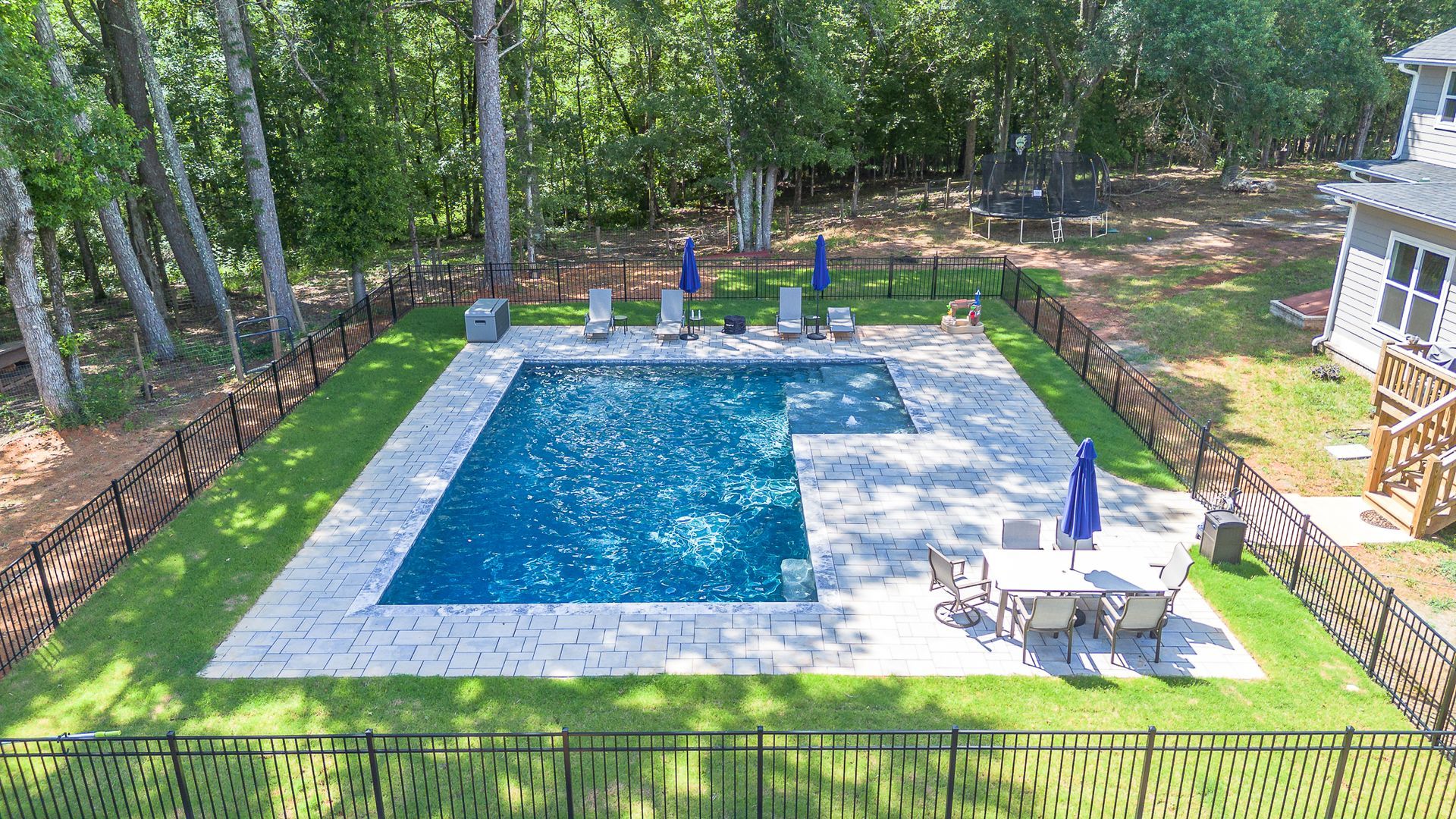 Rectangular pool with patio and dining set, surrounded by fence and green lawn, trees in the background.