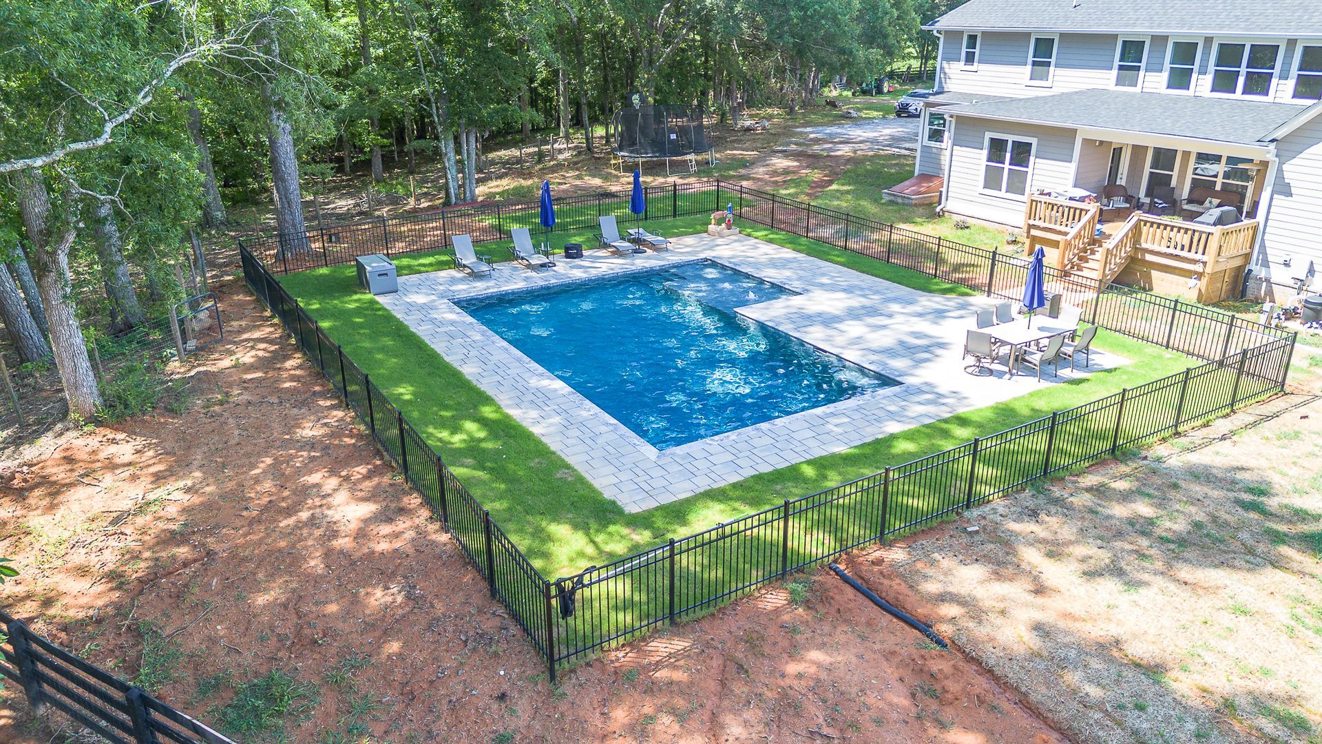 Aerial view of a pool with a house, lawn chairs, and a black fence.