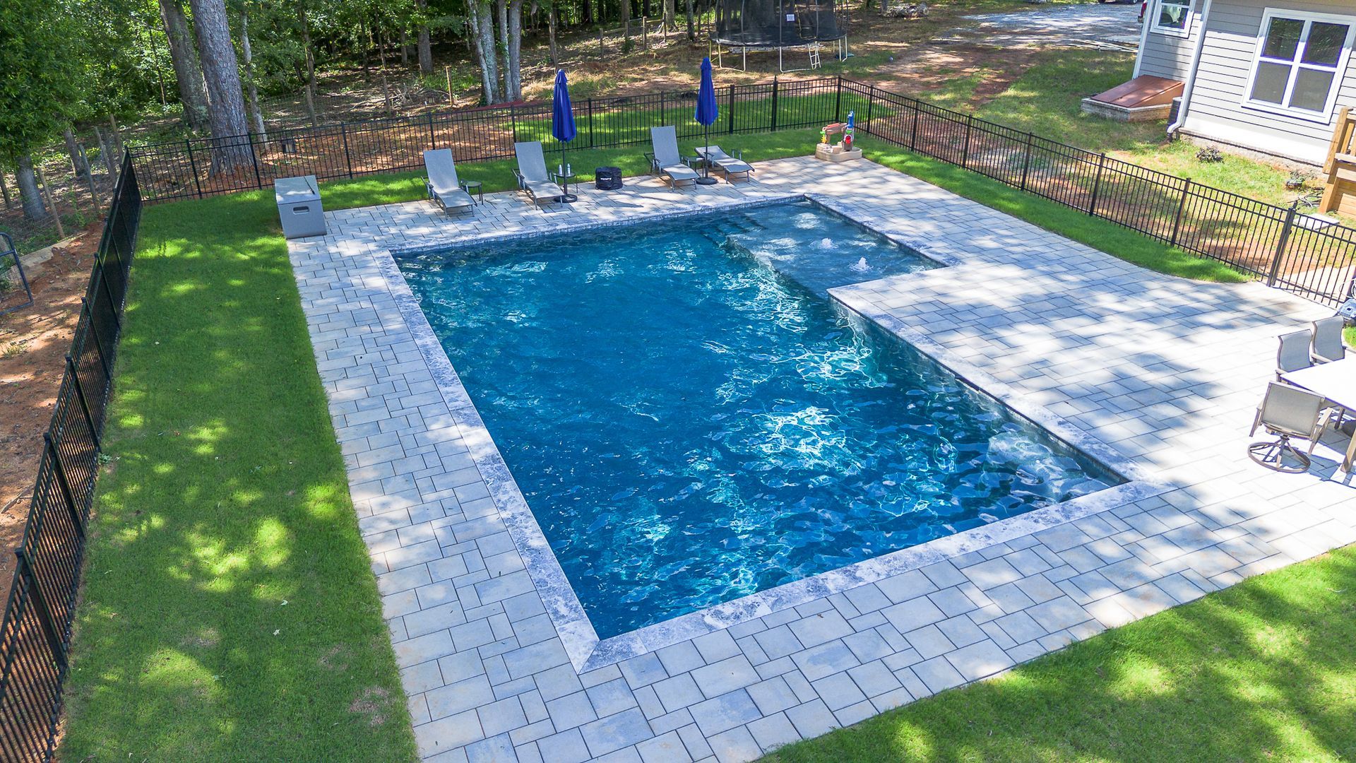 Rectangular swimming pool with paved surround, lounge chairs, and black fence on a green lawn.