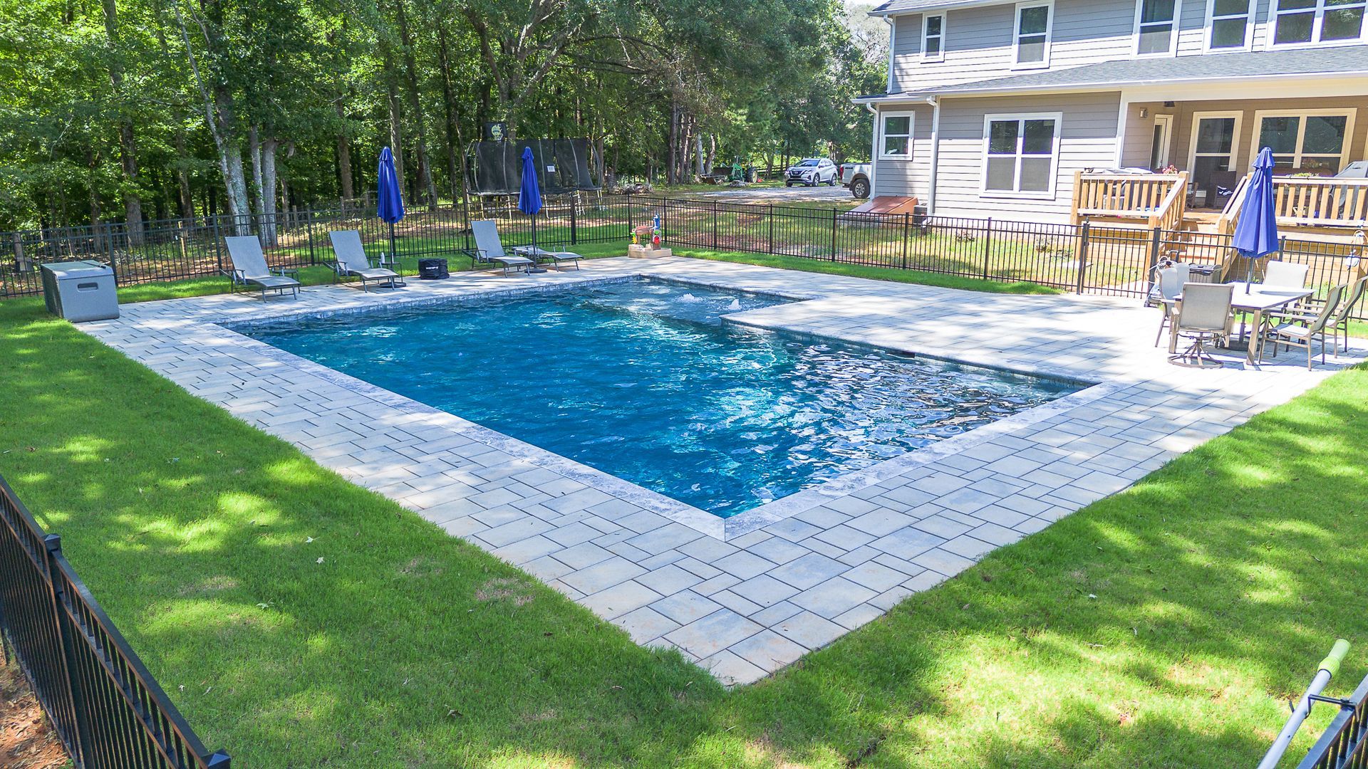 Rectangular pool with blue water, surrounded by gray pavers and green grass, near a house with a deck.