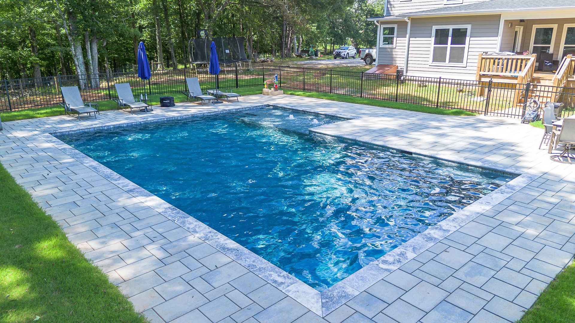 Rectangular swimming pool surrounded by gray pavers and green lawn, near a house with a deck and trees.