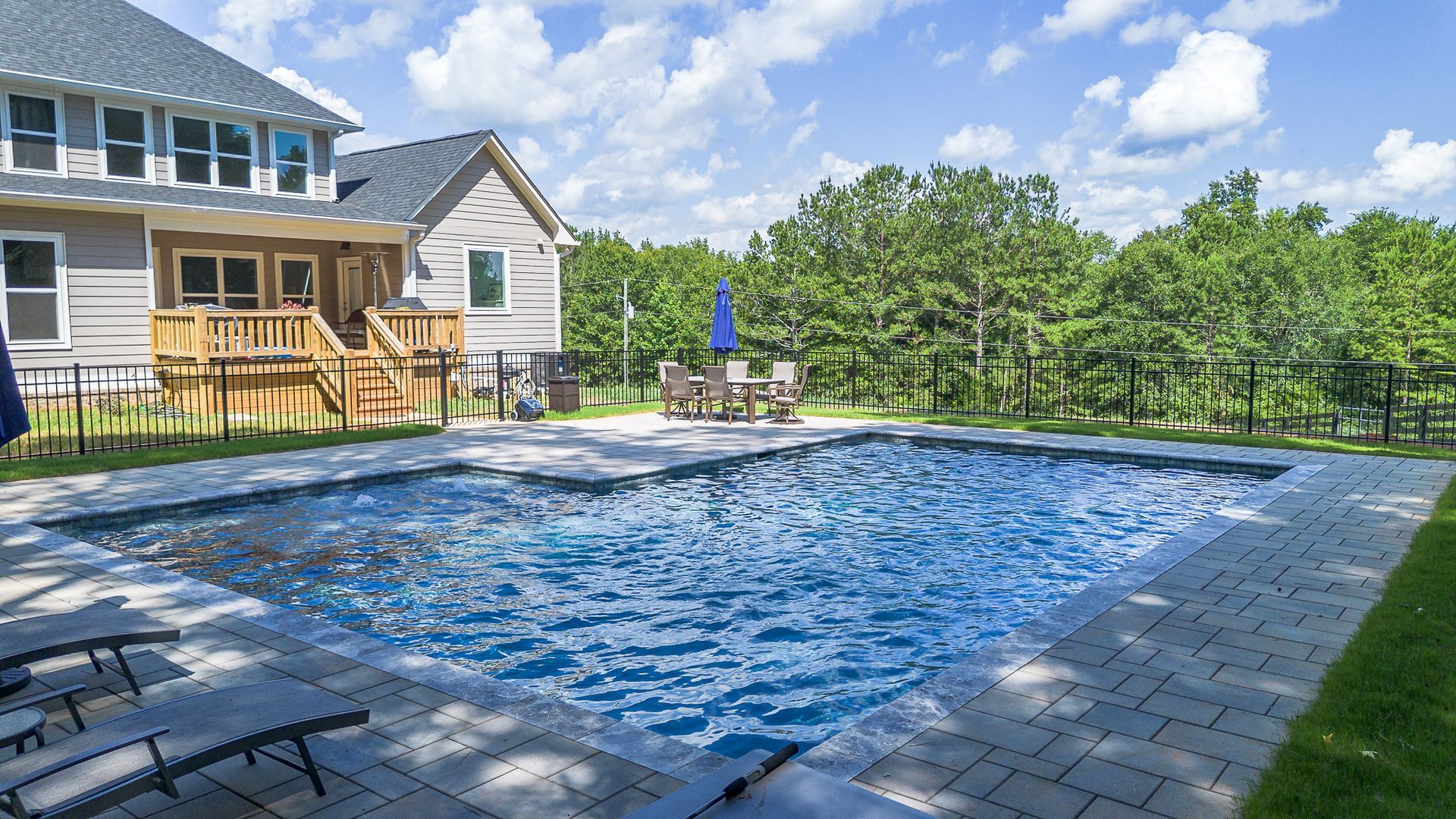 Backyard with a rectangular pool, lounge chairs, and a dining area under a blue umbrella next to a large house.