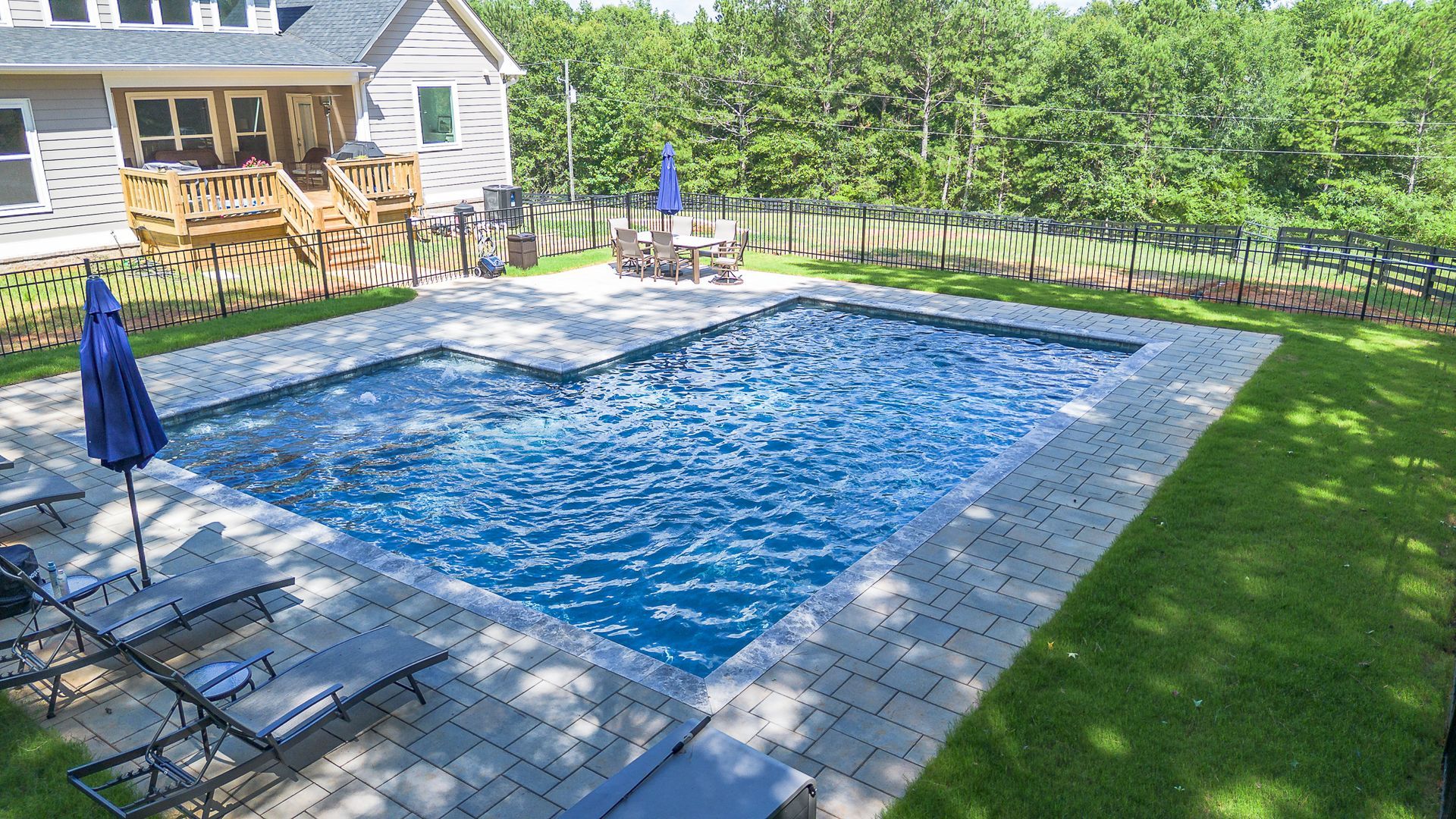 Backyard pool with lounge chairs, blue umbrellas, and a house in the background.