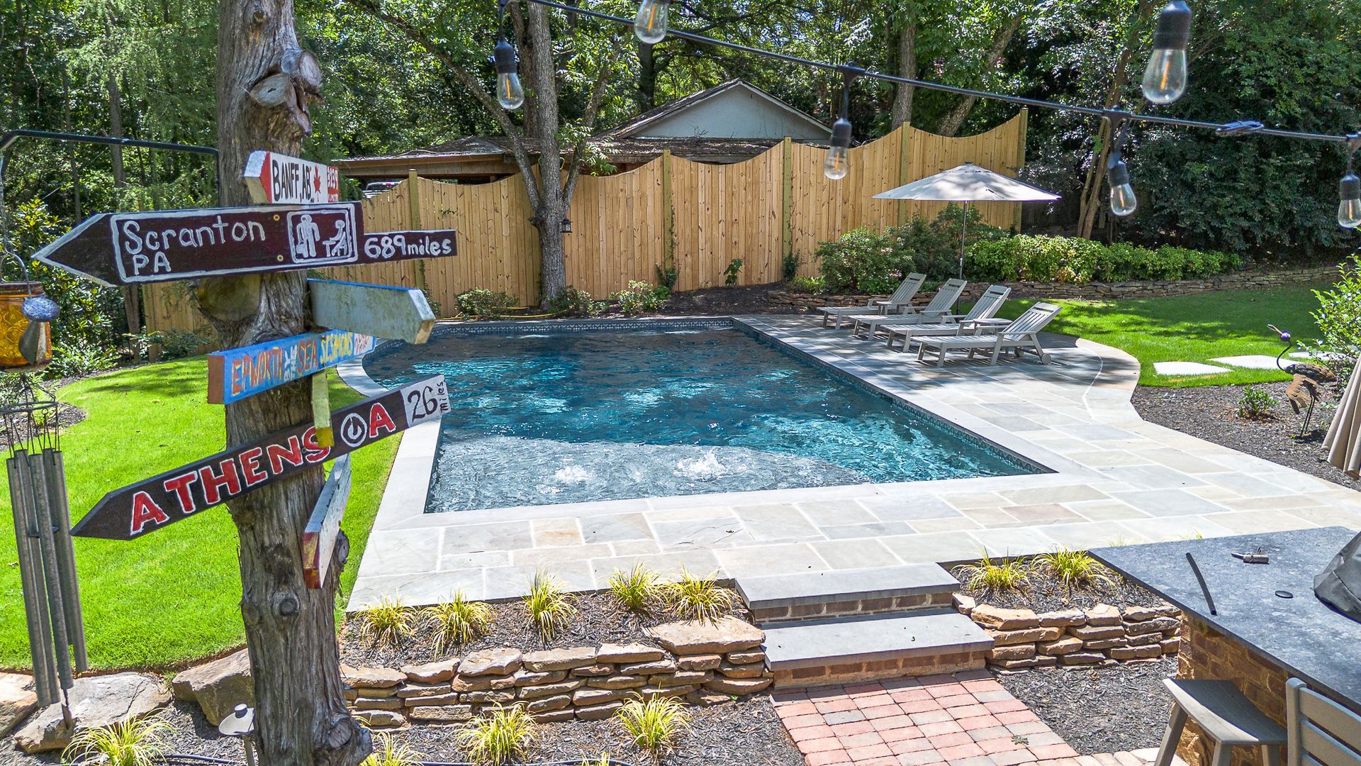 Backyard with pool, wooden fence, string lights, signs, and lounge chairs.