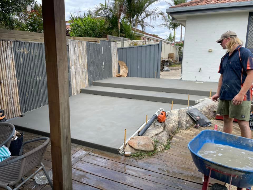 A Man Is Standing Next to A Wheelbarrow Filled with Concrete — J’s Concreting & Carpentry in Bonny Hills, NSW
