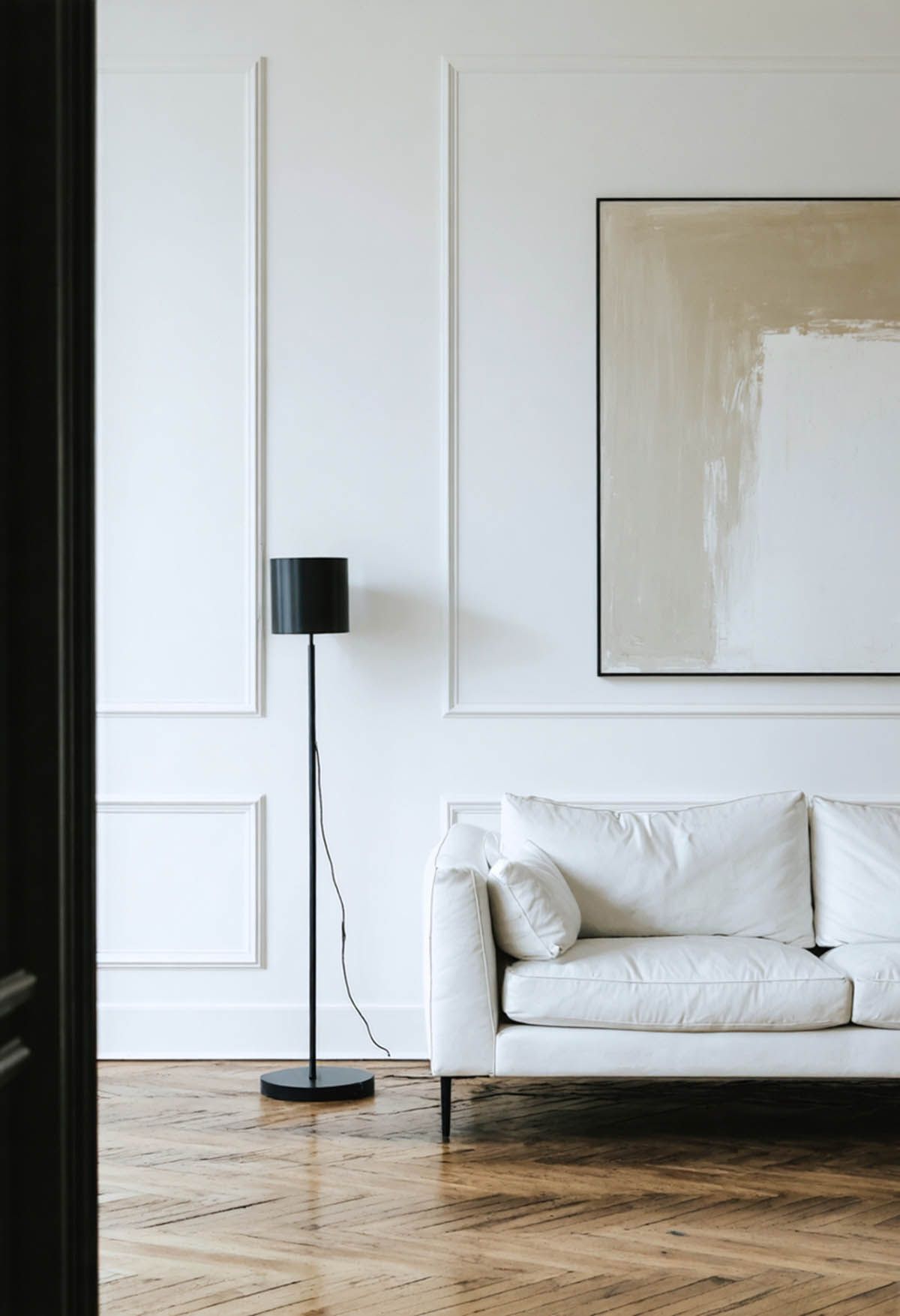 An all white living room with light timber floors, a white sofa and wall paneling. 