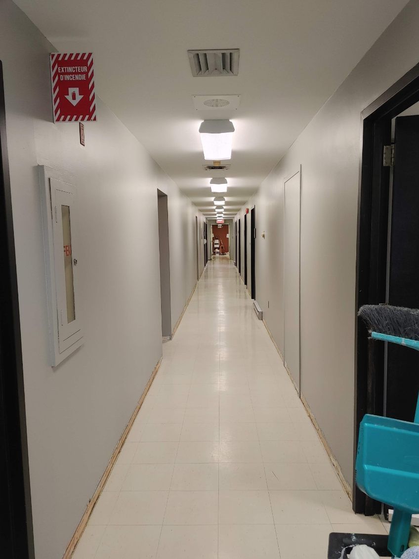 Long couloir blanc avec portes fermées, éclairage au plafond et alarme incendie.