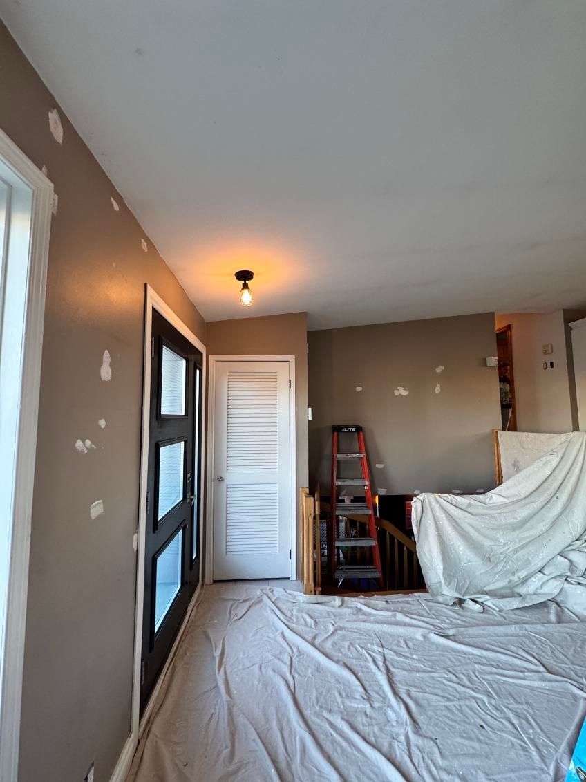Pièce en rénovation, sols recouverts, murs rapiécés, porte noire, porte blanche, échelle et un chiffon.