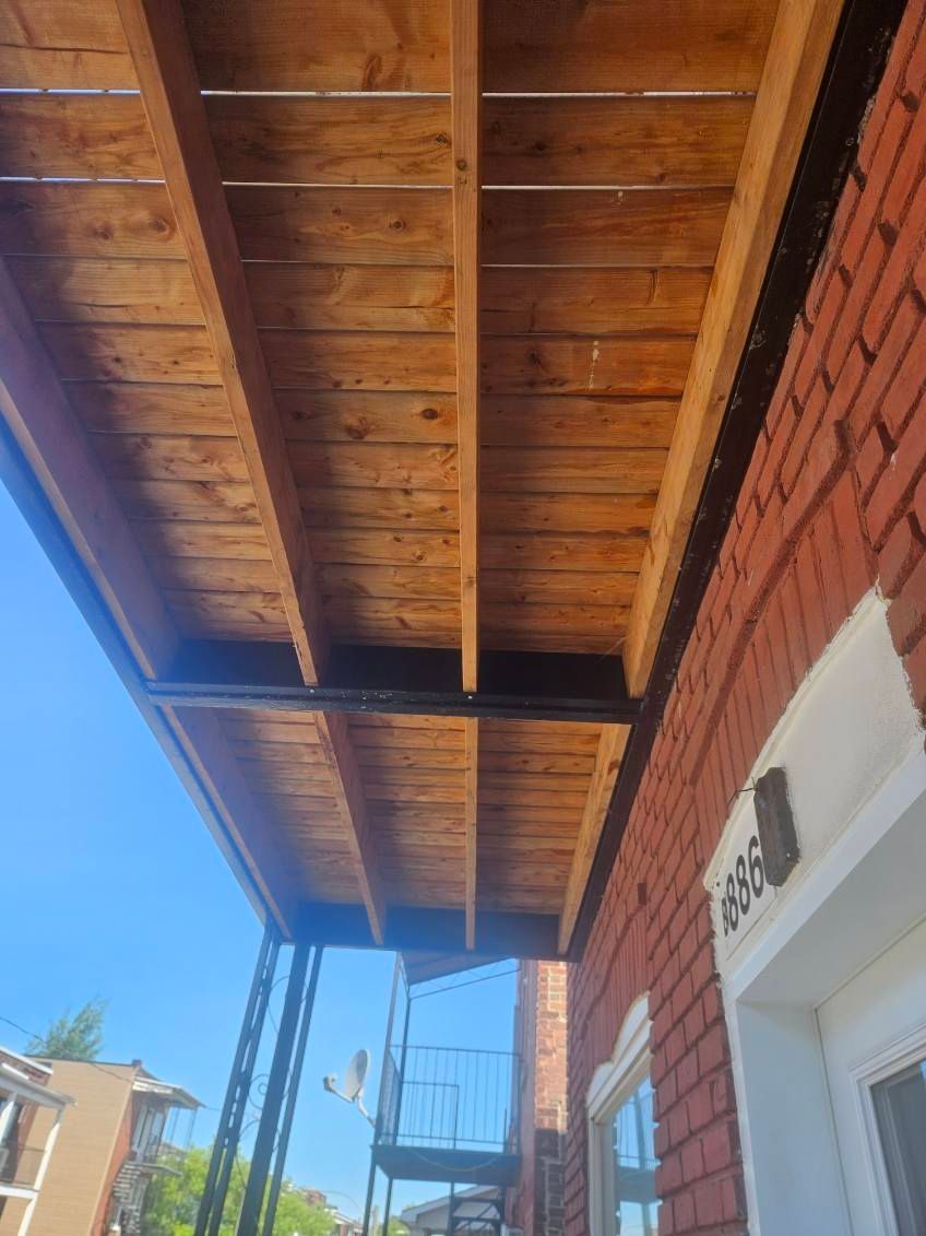 Vue d'un plafond de balcon en bois, soutenu par des poutres métalliques, fixé à un bâtiment en briques.