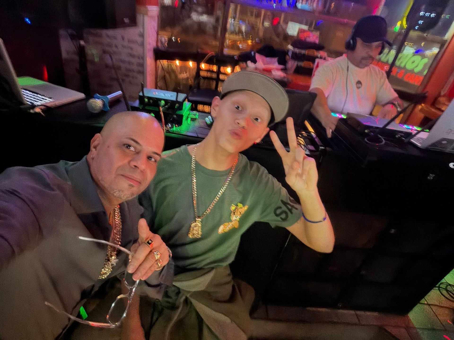 Two people posing for a selfie in a dimly lit nightclub, with a DJ booth and neon lights behind them.