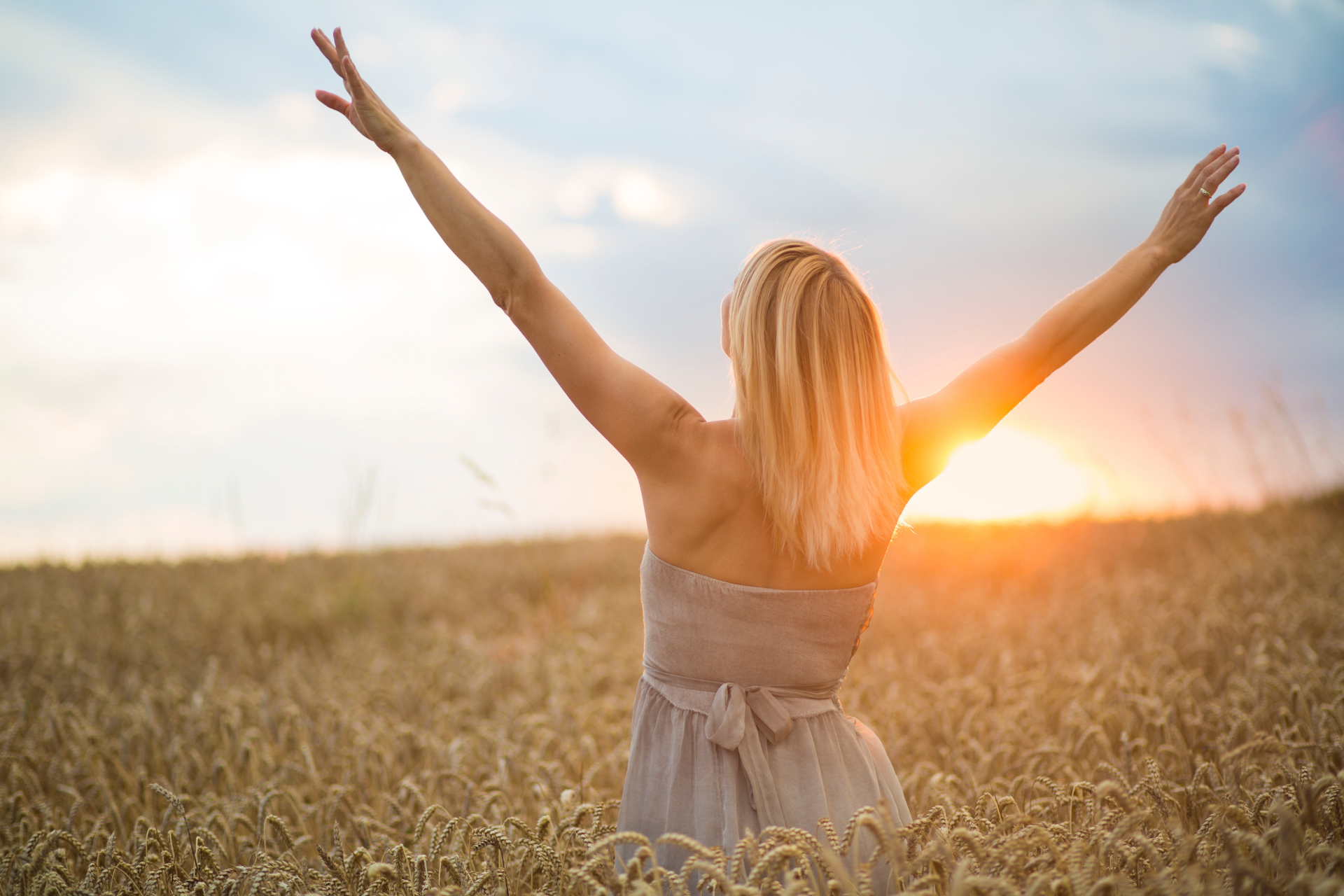 A person stands in a sunlit field of wheat with their arms raised toward the sky at sunset.