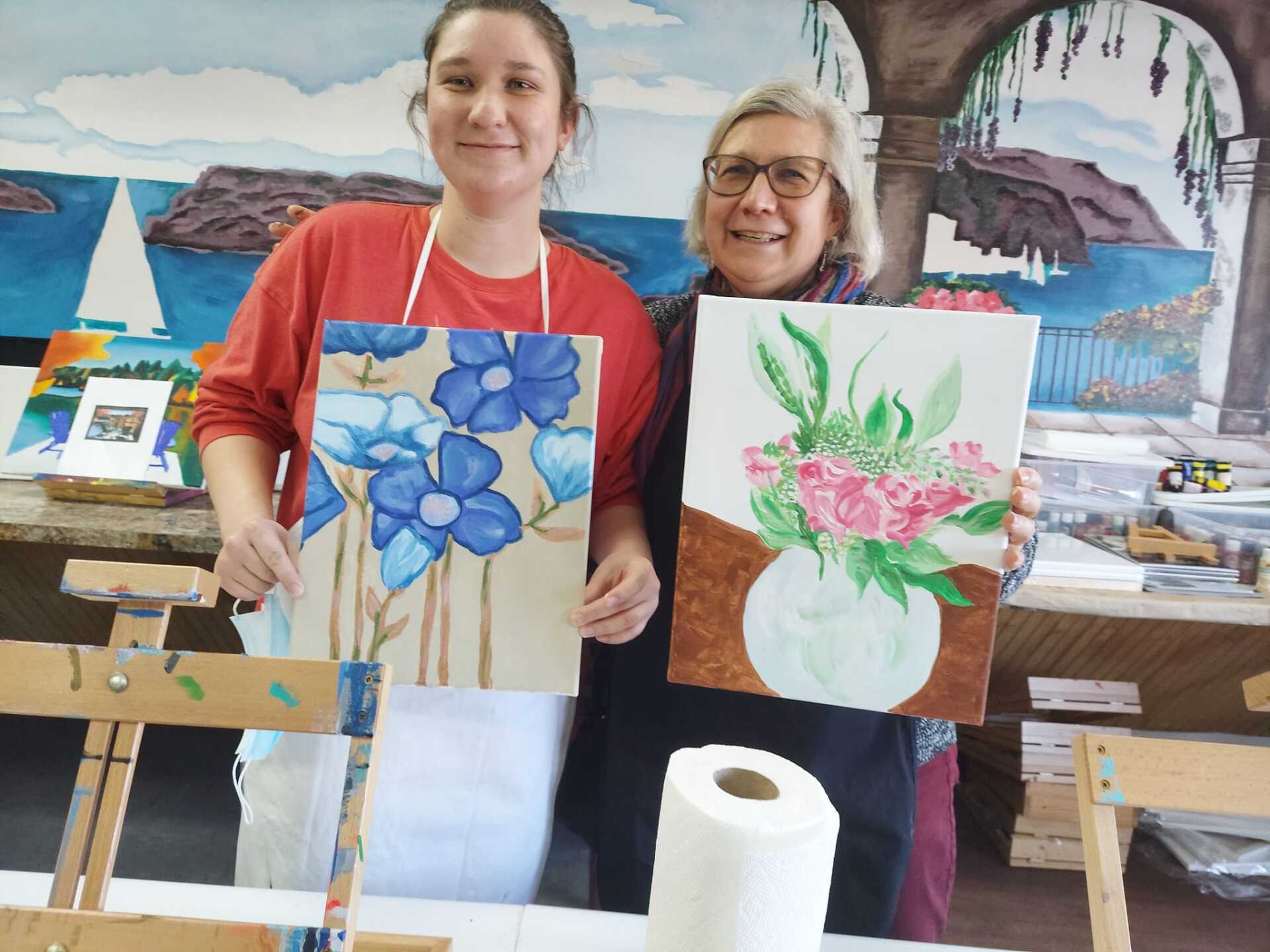 Two women are standing next to each other holding paintings of flowers.