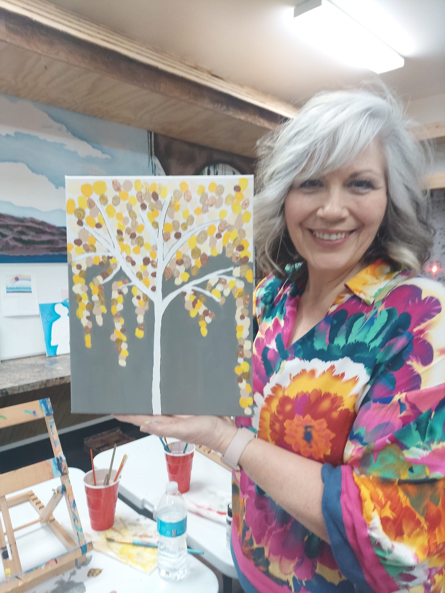 A woman is holding a painting of a tree with yellow leaves.
