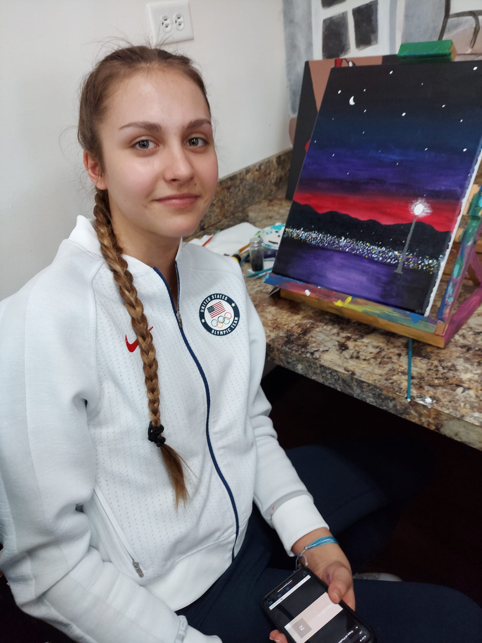 A young woman is sitting in front of a painting on an easel.