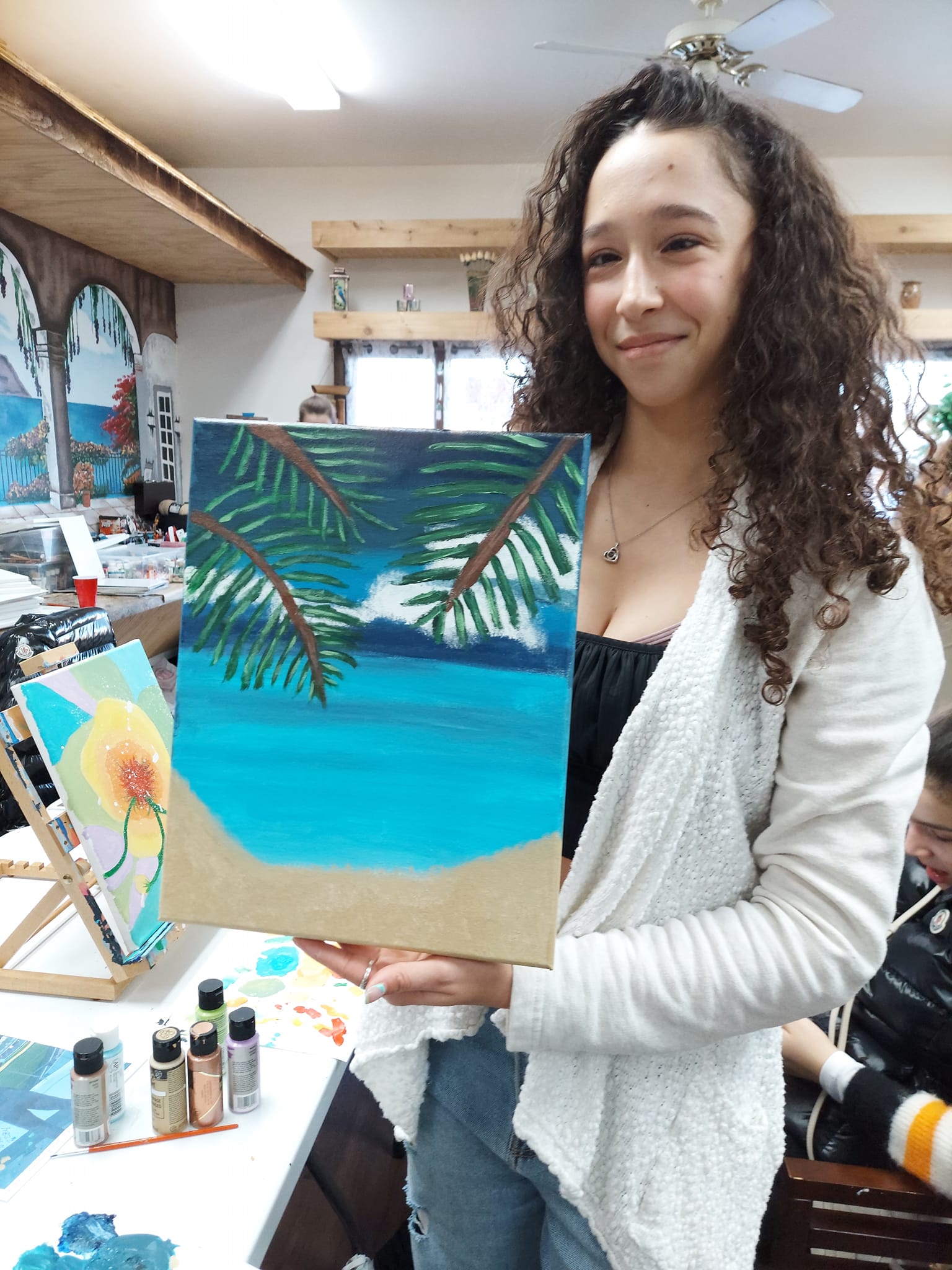 A woman is holding a painting of a beach and palm trees.
