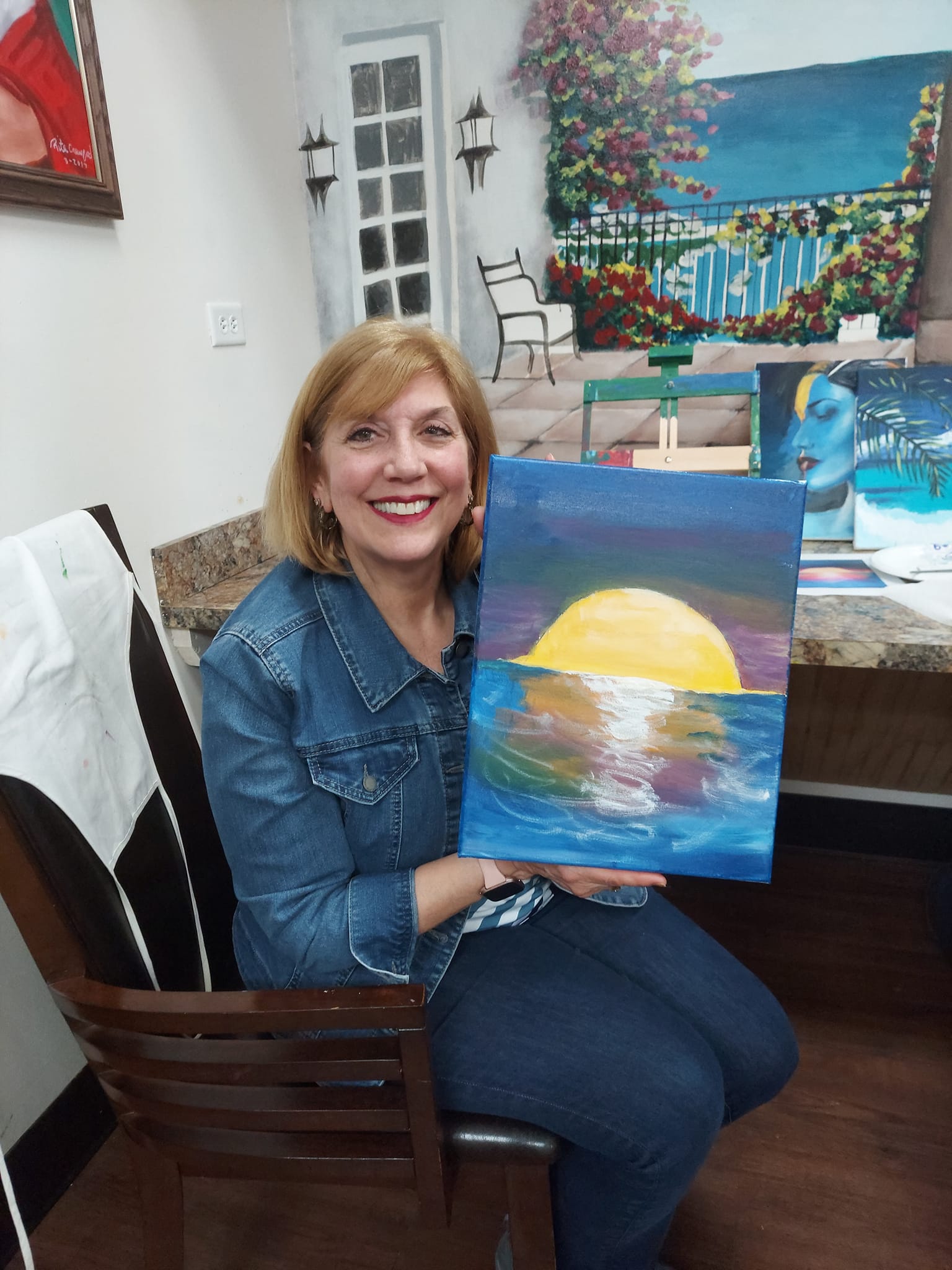 A woman is sitting in a chair holding a painting of a sunset over the ocean.