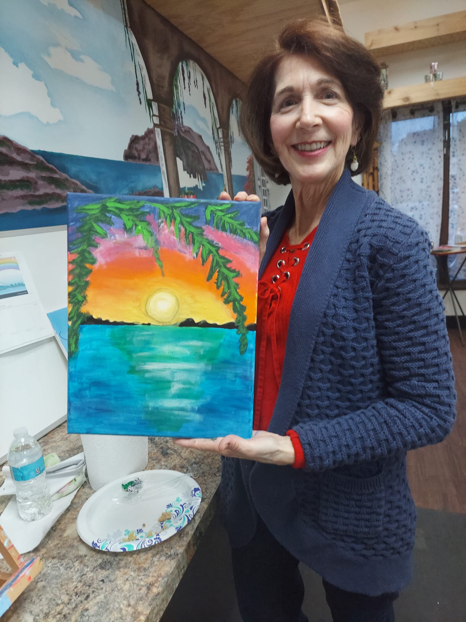 A woman is holding a painting of a sunset over the ocean.