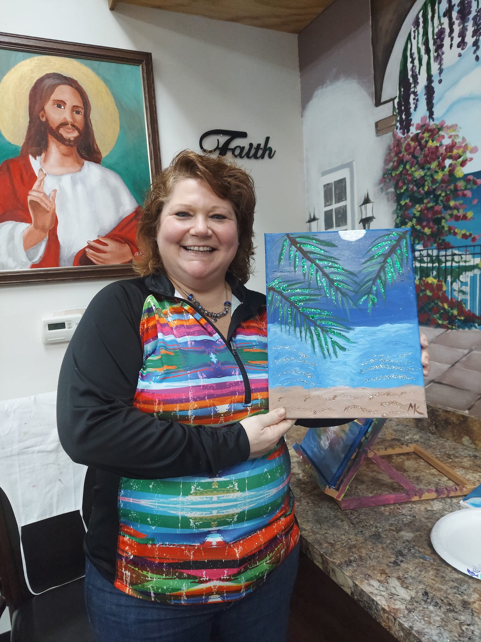 A woman is holding a painting of a beach in front of a painting of jesus.