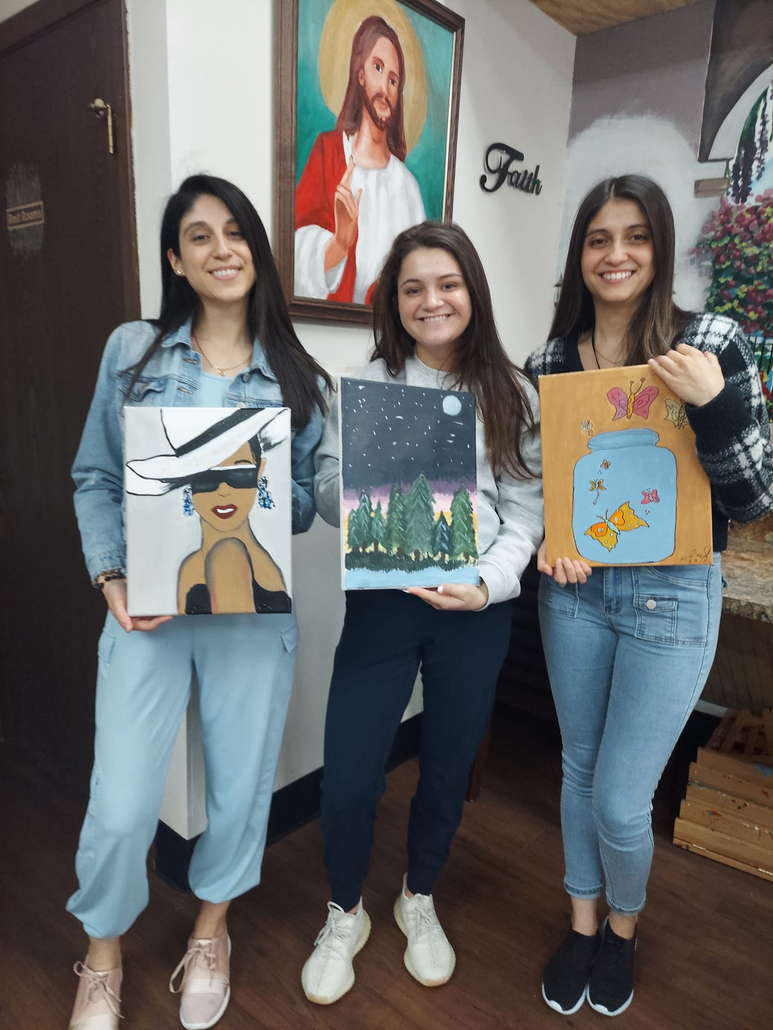 Three women are standing next to each other holding paintings in front of a painting of jesus.