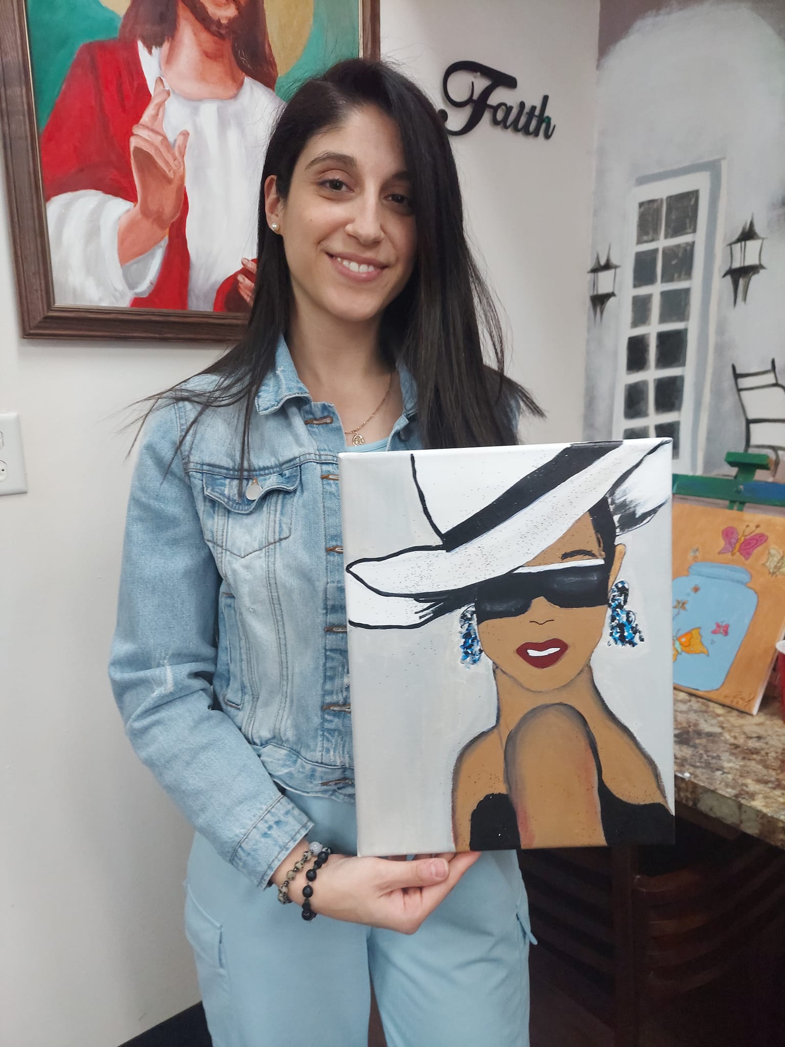 A woman is holding a painting of a woman wearing a hat and sunglasses.