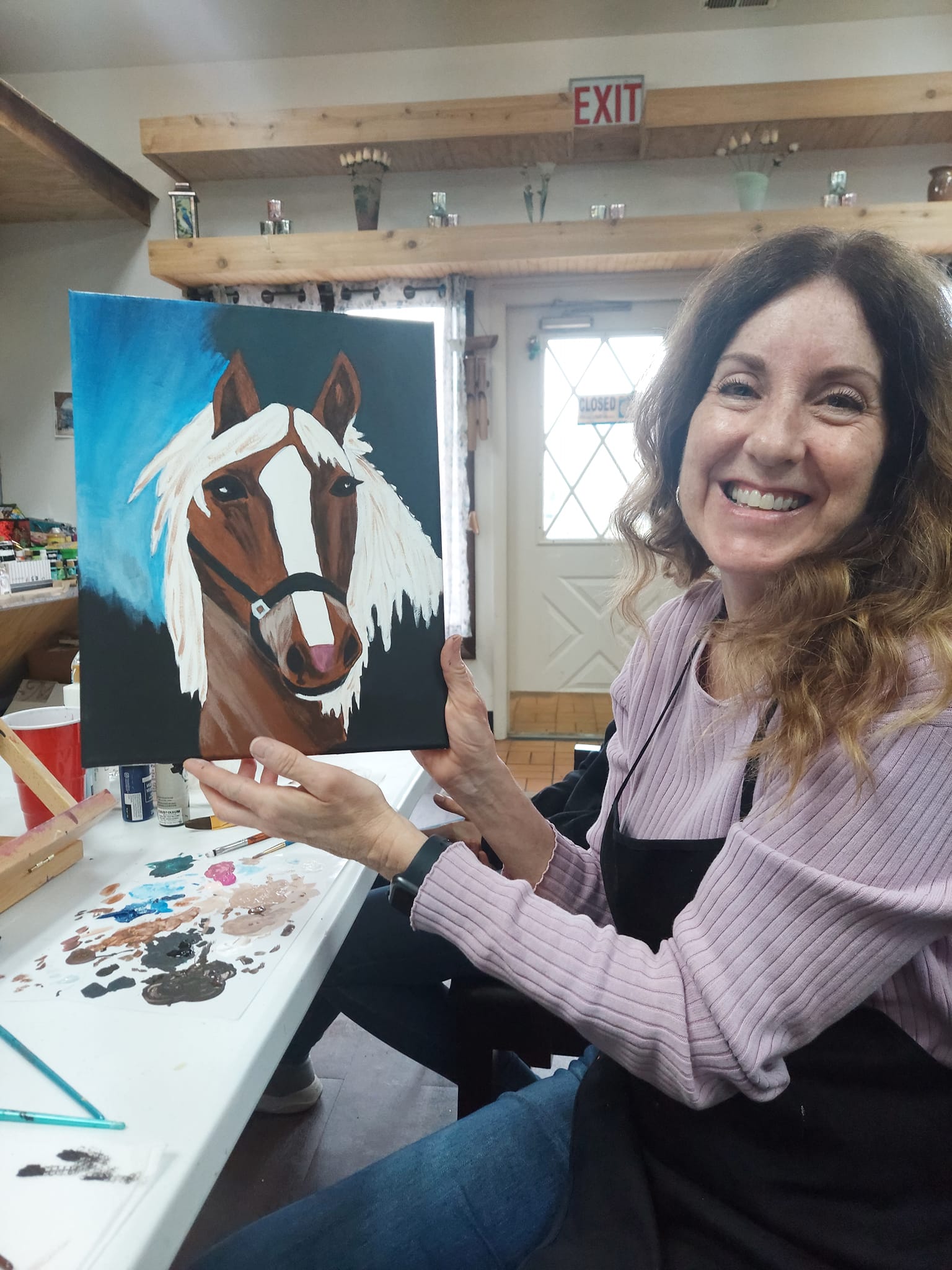 A woman is sitting at a table holding a painting of a horse.