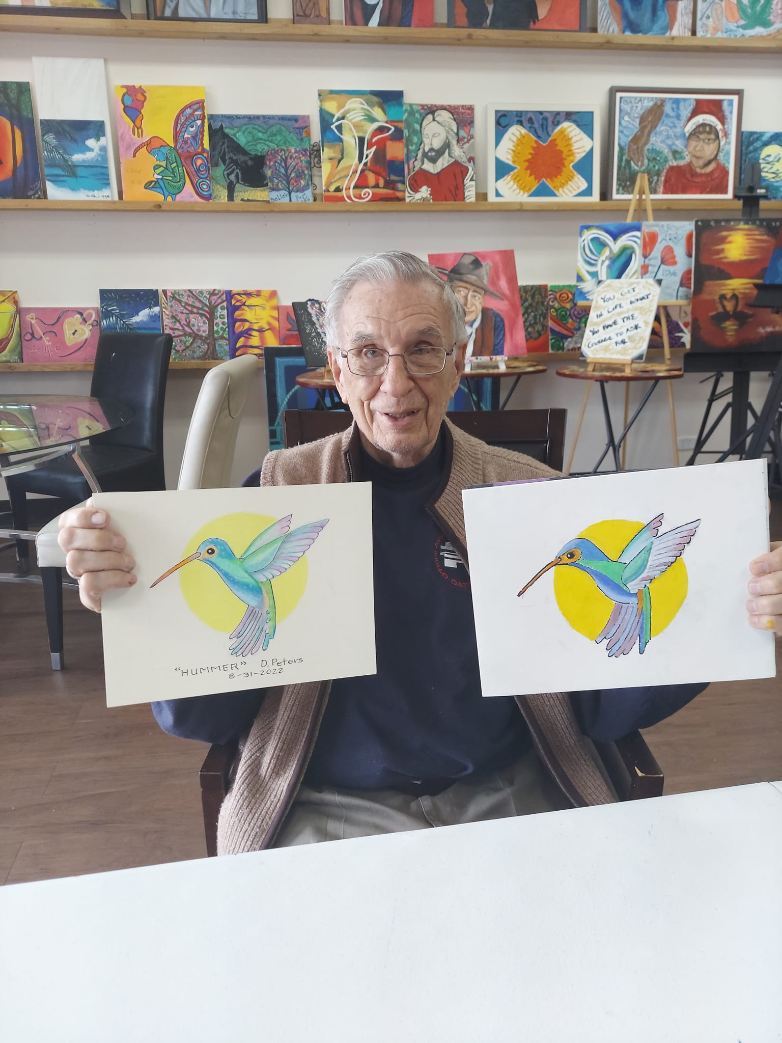 An elderly man is holding two paintings of hummingbirds
