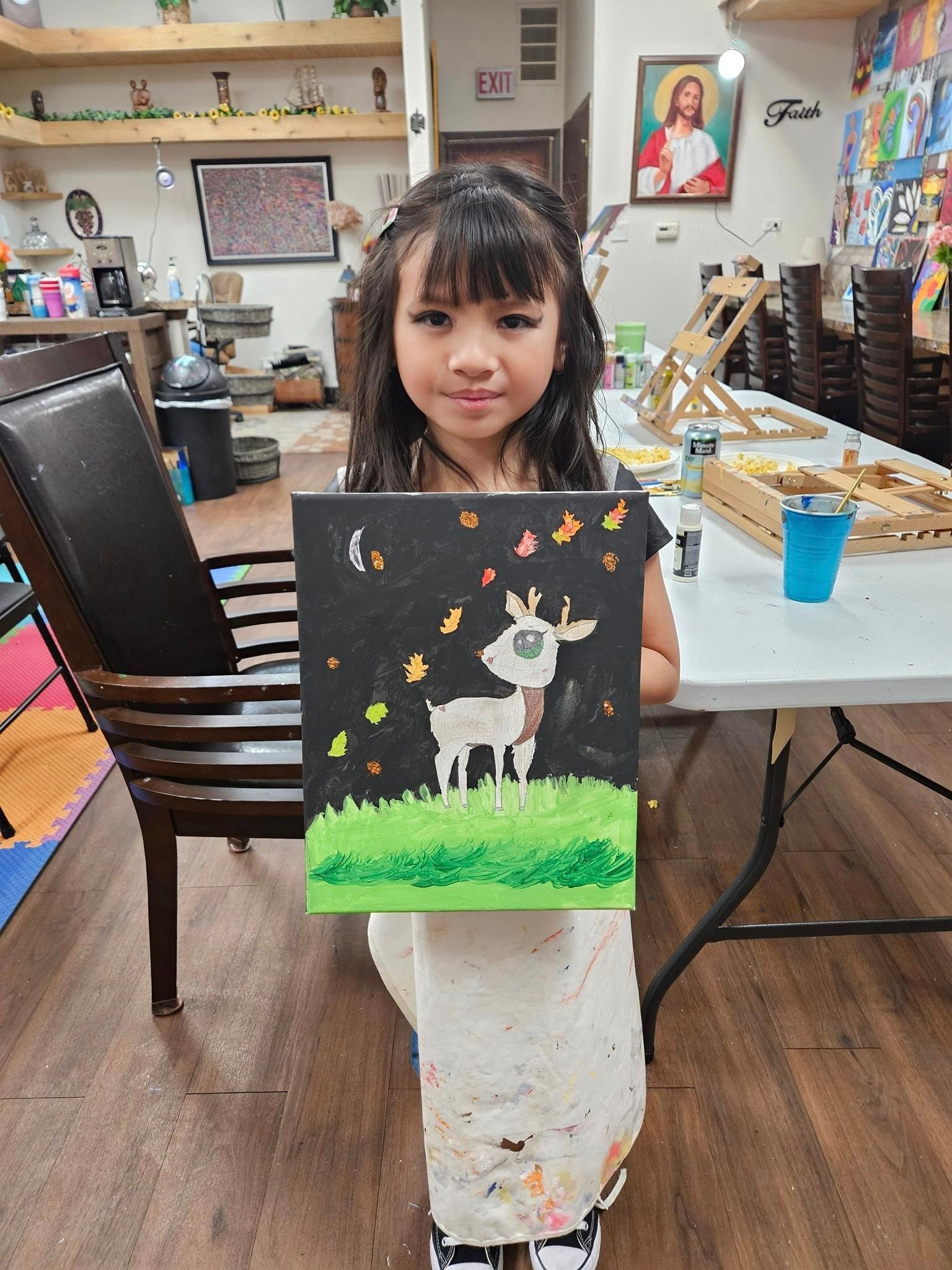 A little girl is holding a painting of a goat in a field.