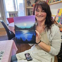 A woman is sitting at a table holding a painting of a lake.