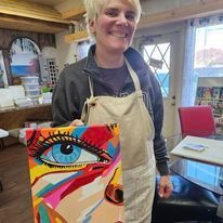 A woman in an apron is holding a colorful painting of a woman 's eye.
