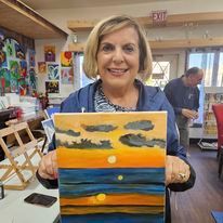 A woman is holding a painting of a sunset over the ocean.