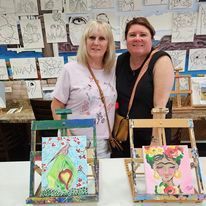 Two women are standing next to each other in front of a table with paintings on it.