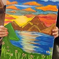 A person is holding a painting of a sunset over a lake with mountains in the background.