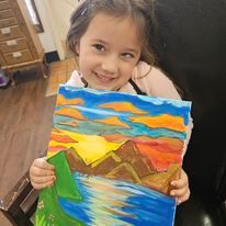 A little girl is holding a painting of a sunset over a lake.