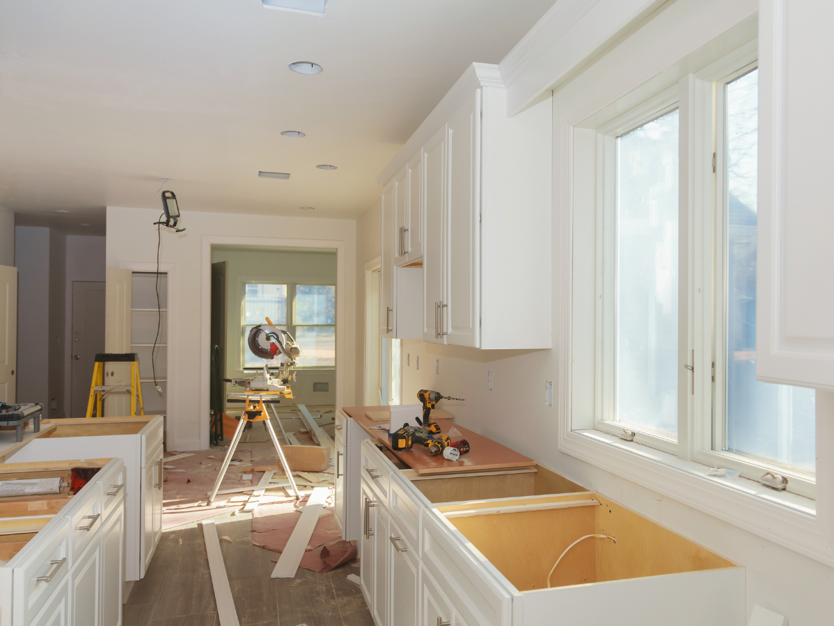 Bright kitchen under renovation with white cabinets, sunlit windows, and tools on the floor