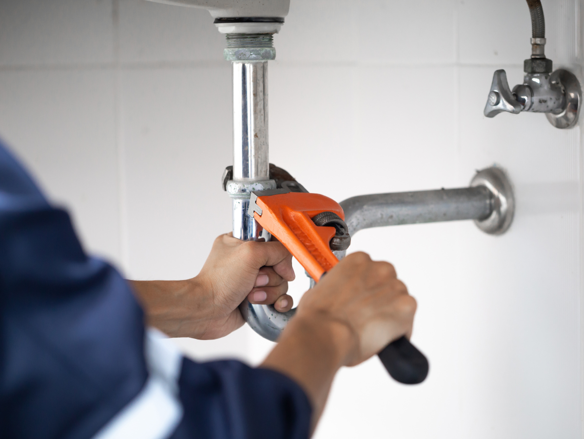 Plumber tightening a sink pipe under a bathroom sink with an orange pipe wrench