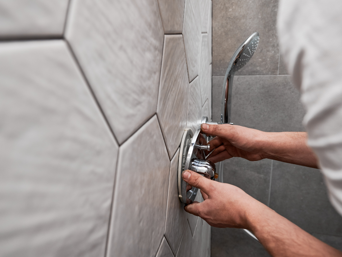 Hands adjusting a chrome shower control in a tiled bathroom