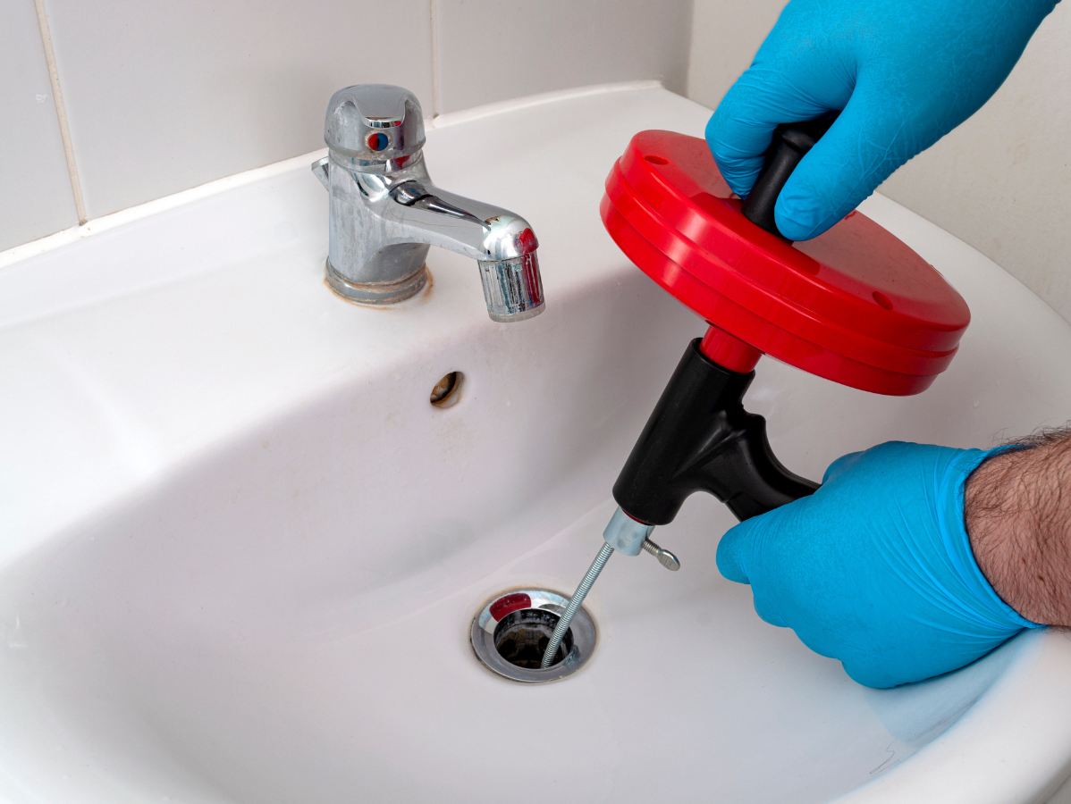 Gloved hands using a red drain auger in a white bathroom sink