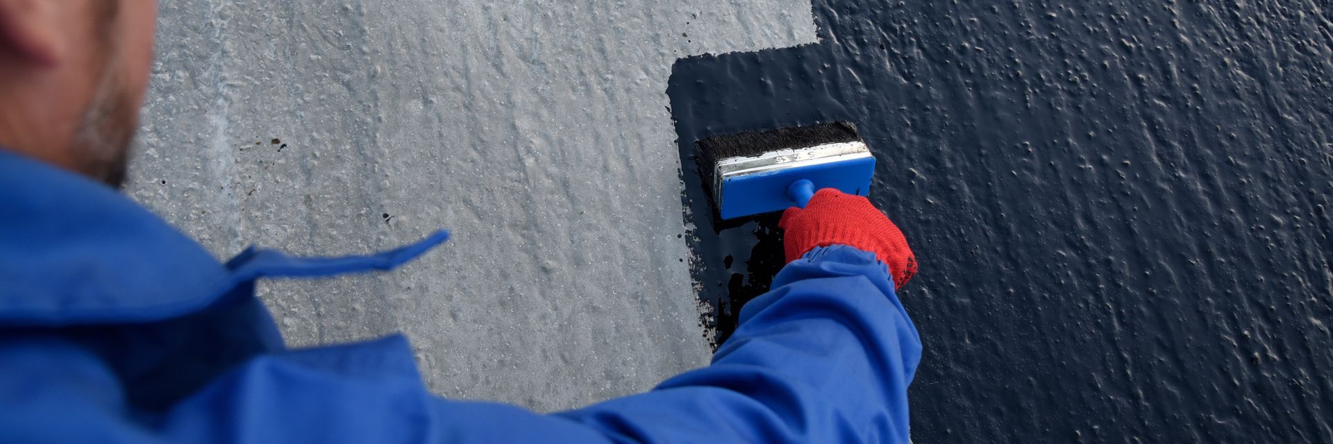 Gloved hand applying black coating to a rough wall with a brush.