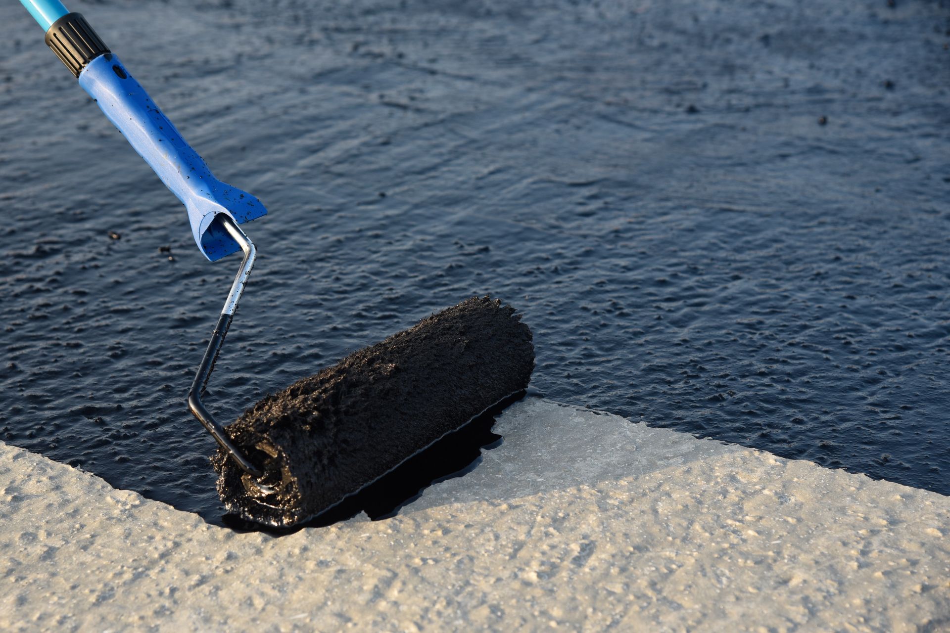 A blue-handled tool applying black sealant along a concrete edge by water