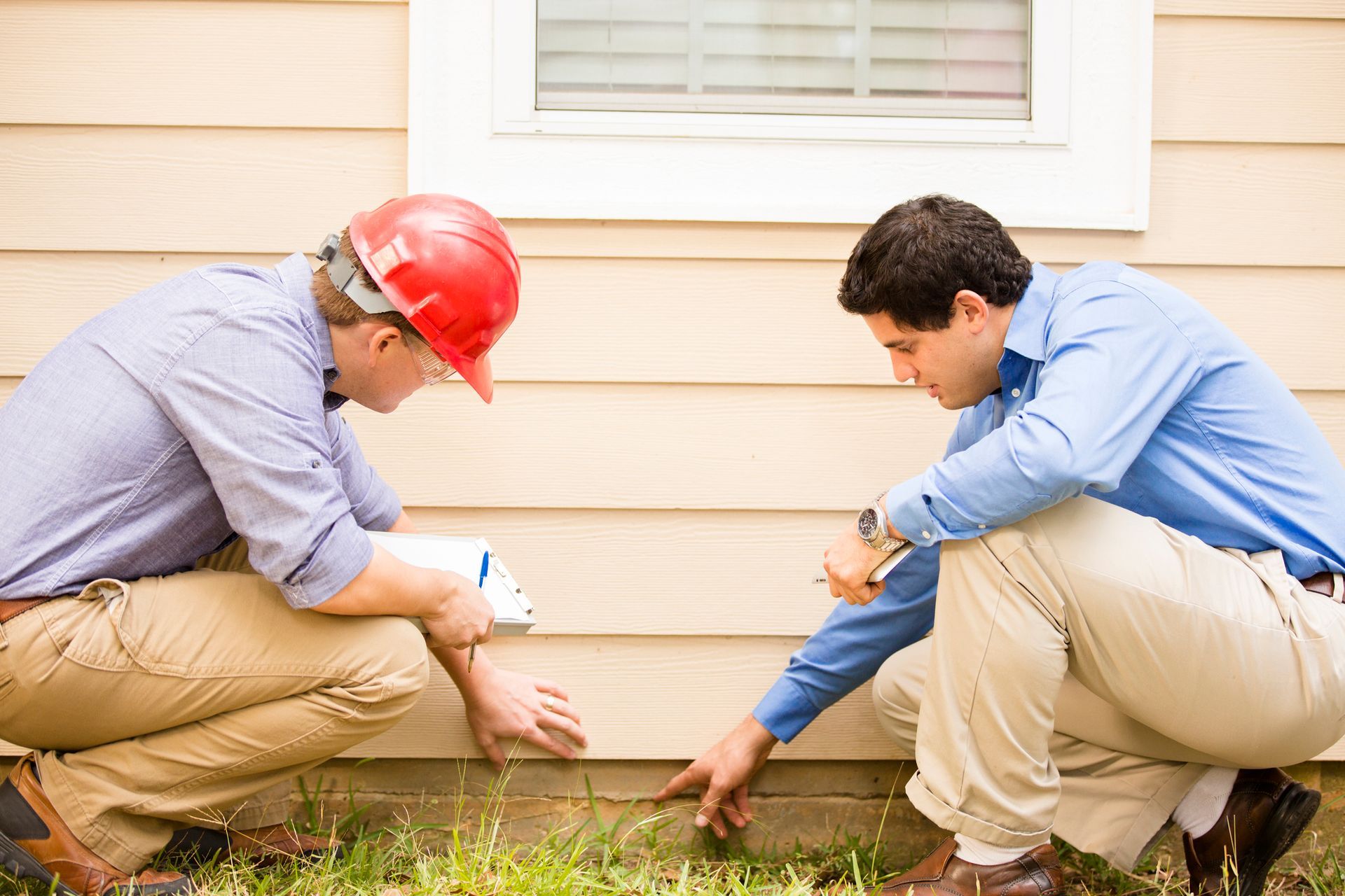 Two people crouching beside a house, examining the foundation near the exterior siding.