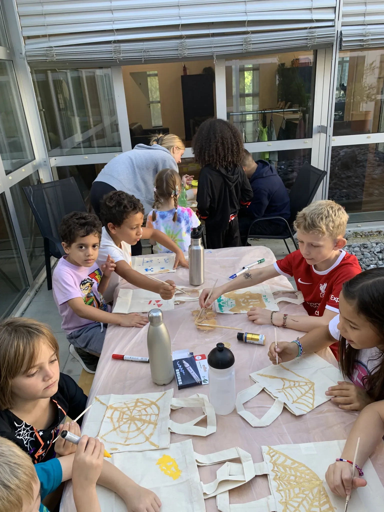 A group of children sit around a table in a sunroom, painting designs onto canvas tote bags.