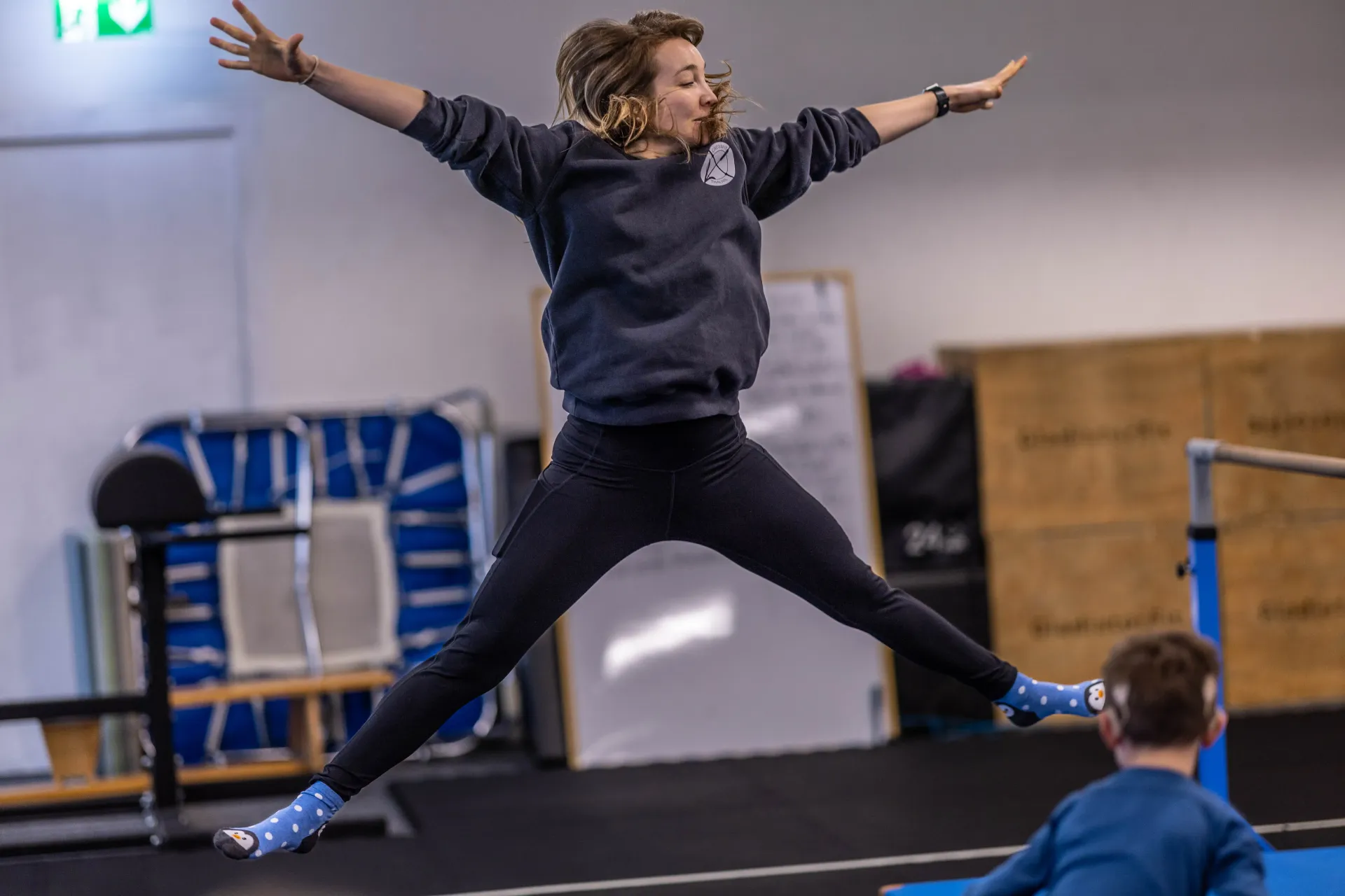 A person performs a star jump in an indoor gym in Winterthur with a child watching in the foreground.