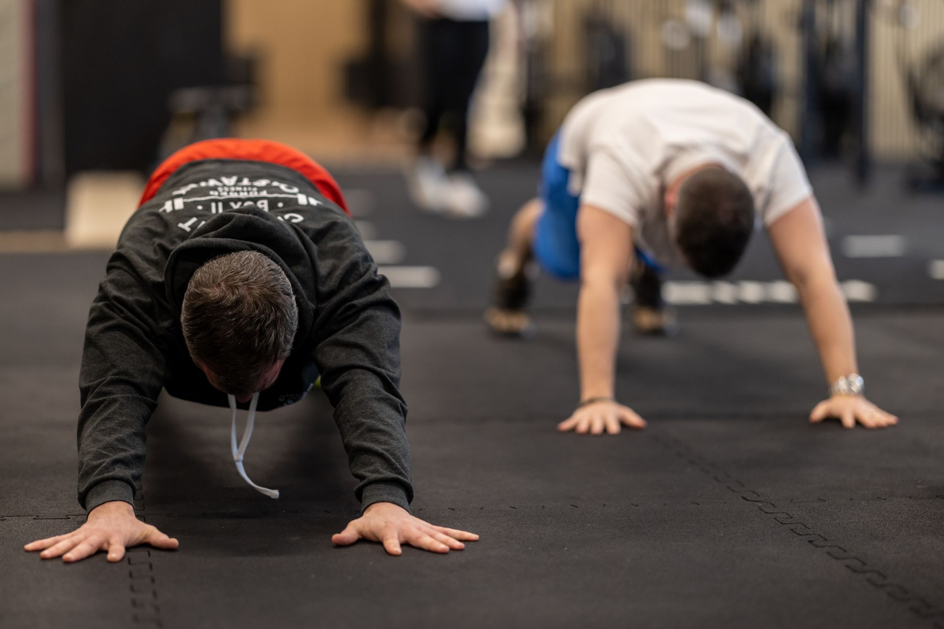 Two people in a gym in Winterthur perform plank exercises on black floor mats.